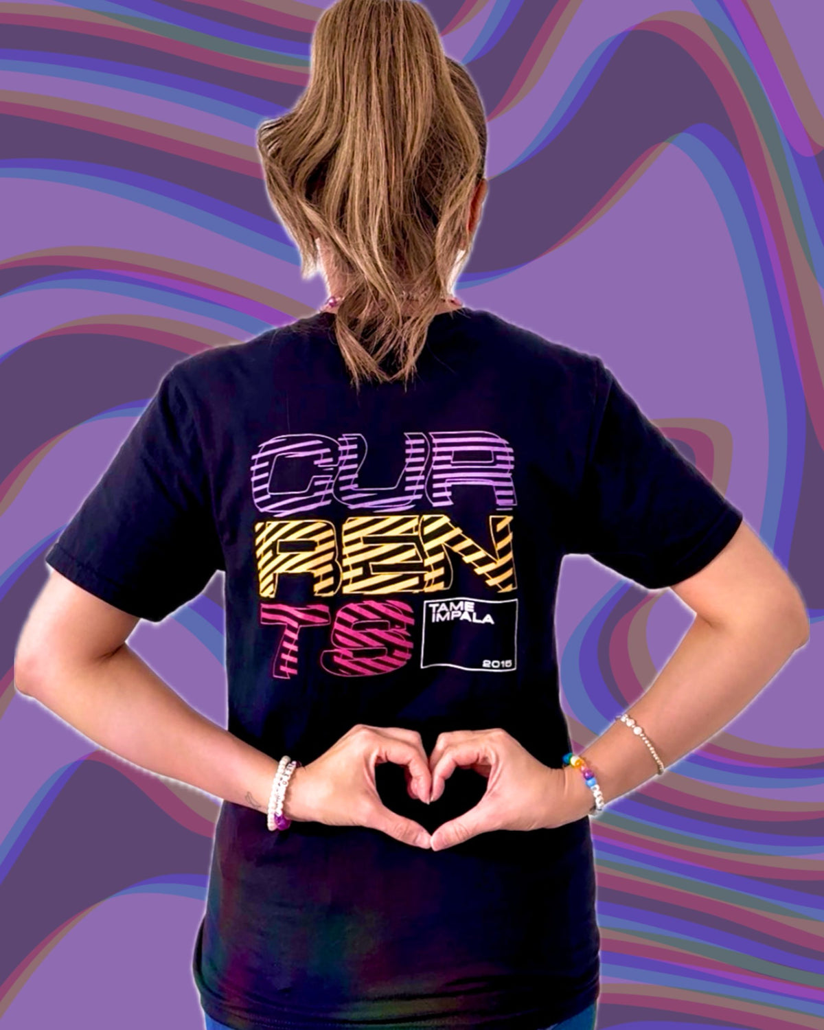 A woman shown from behind wearing a black graphic t-shirt with the words 'CURRENTS' in colorful retro striped and zebra-print lettering. She is posing with her hands forming a heart shape at her back against a purple and pink wavy background.