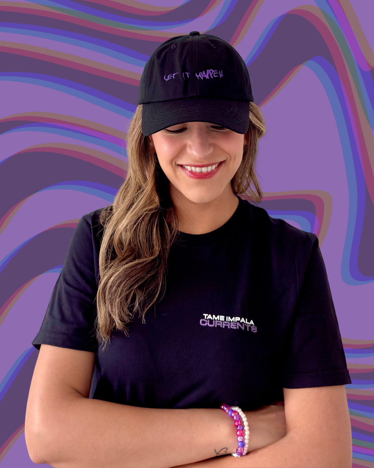 Model is wearing a black t-shirt with "TAME IMPALA" in small white letters with "CURRENTS" underneath it in purple text. Text is located on the left chest design. Model is also wearing a black cap with purple, handwritten embroidered text that says "Let It Happen"