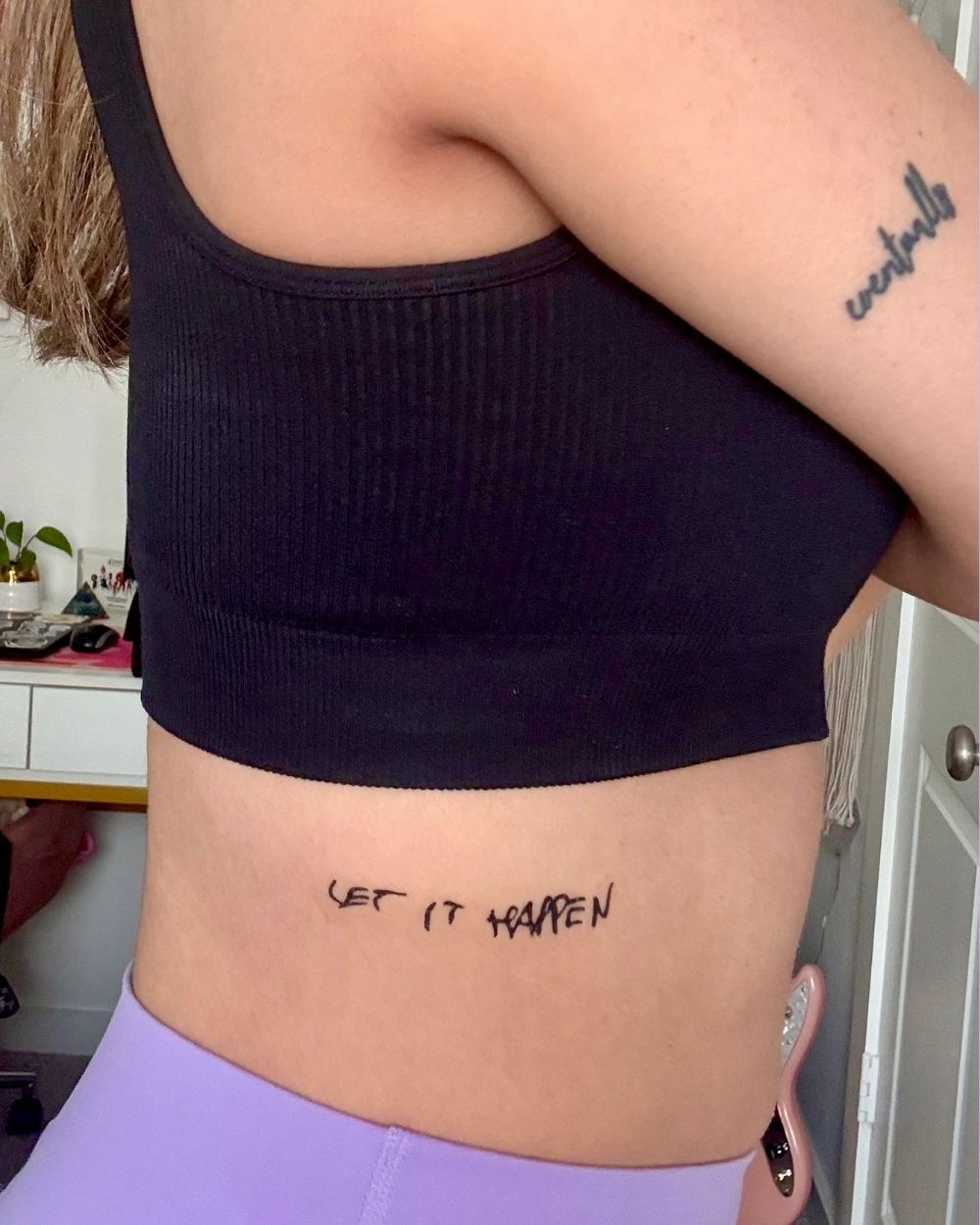 A close-up of a person's side ribs with a small black script tattoo reading 'LET IT HAPPEN' and another tattoo is visible on the upper arm.