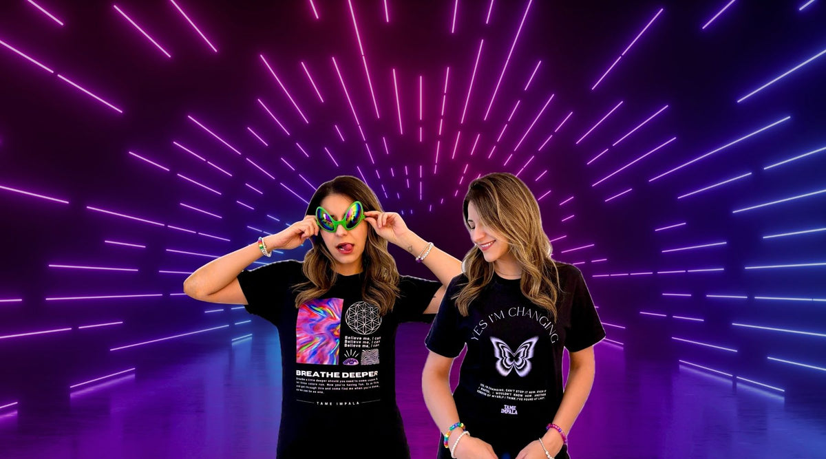 Two women modeling black graphic t-shirts in front of a neon tunnel backdrop. The woman on the left wears green sunglasses and a t-shirt with a colorful graphic and the text 'BREATHE DEEPER'. The one on the right wears a shirt that says "Yes I'm Changing" with a light lavender butterfly graphic.