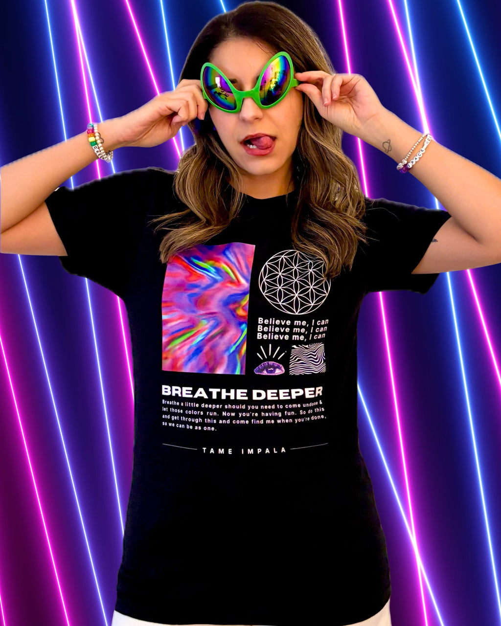 A woman wearing a black Tame Impala T-shirt with the words "BREATHE DEEPER", a colorful graphic print and white shorts, posing in front of a neon purple and pink light background. She is holding up green alien sunglasses.