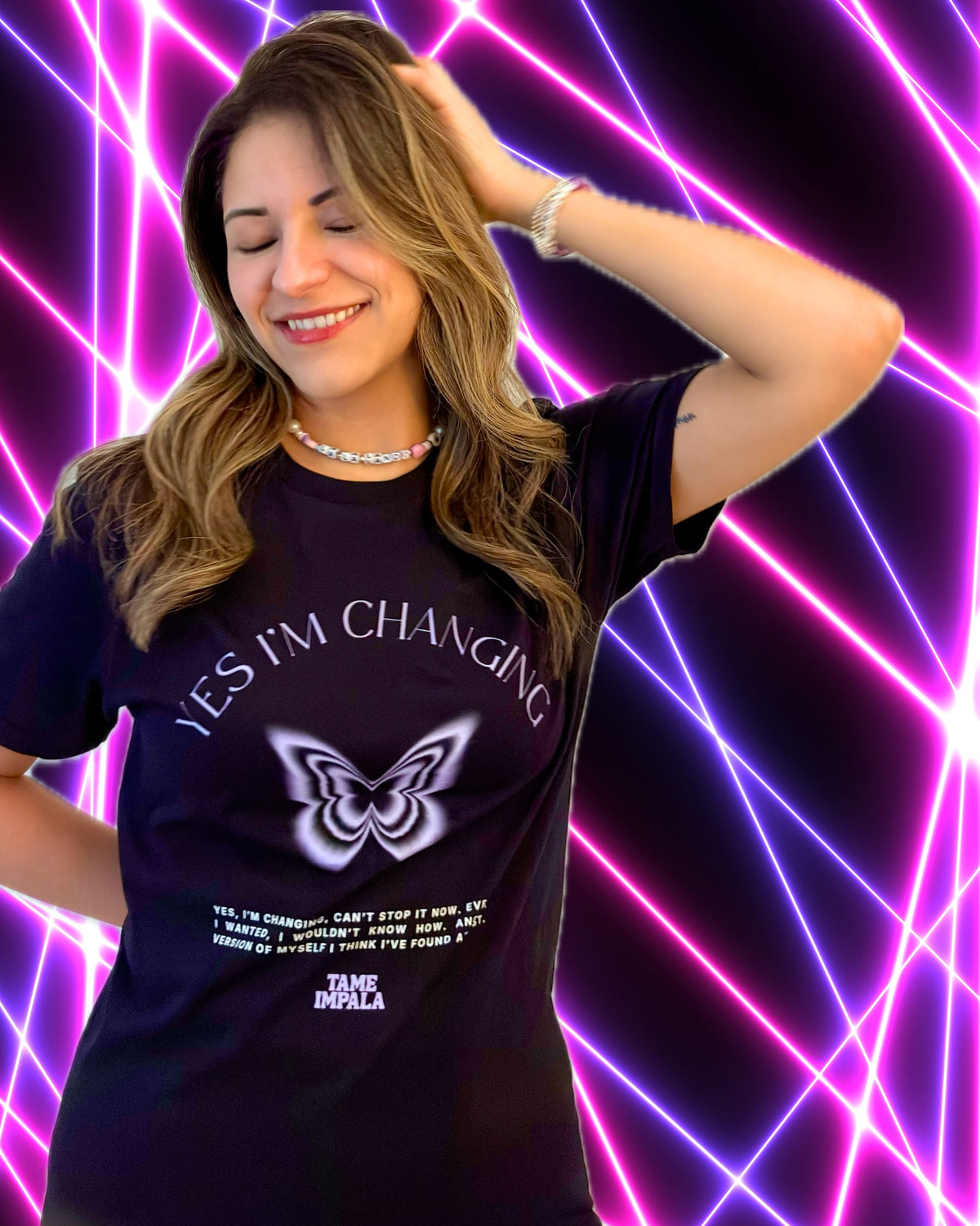 A woman wearing a black graphic t-shirt with a light lavender butterfly design and the text 'YES I'M CHANGING' and lyrics from the Tame Impala song, posed against a vibrant purple and pink neon background.